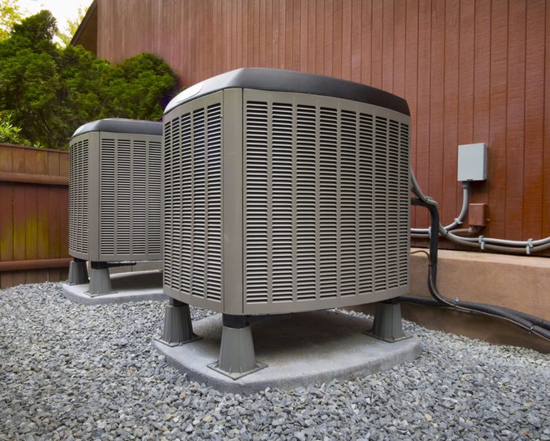 two large outdoor HVAC units next to a wooden wall of a home