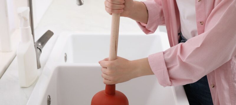 a woman using a plunger on the drain of a kitchen sink