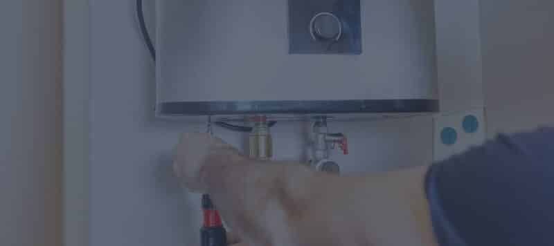 A person's hand uses a screwdriver to work on the plumbing and wiring at the bottom of a metallic tankless water heater